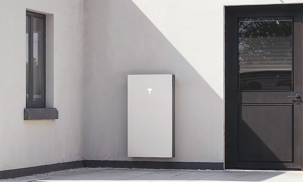 west-sussex-powerwall-installers A Tesla Powerwall 3 battery unit installed on a white exterior wall in direct sunlight at a West Sussex property